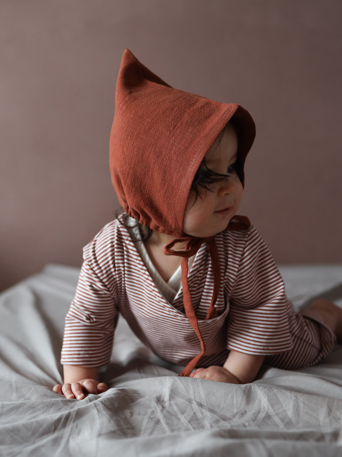 Ein Baby trägt den Wickel Overall Baumwolle - Ringel Ziegel, einen gestreiften Einteiler mit Wickelausschnitt, während es auf einer weichen, hellgrauen Unterlage krabbelt, ideal für warme Tage und handgemacht in Berlin.