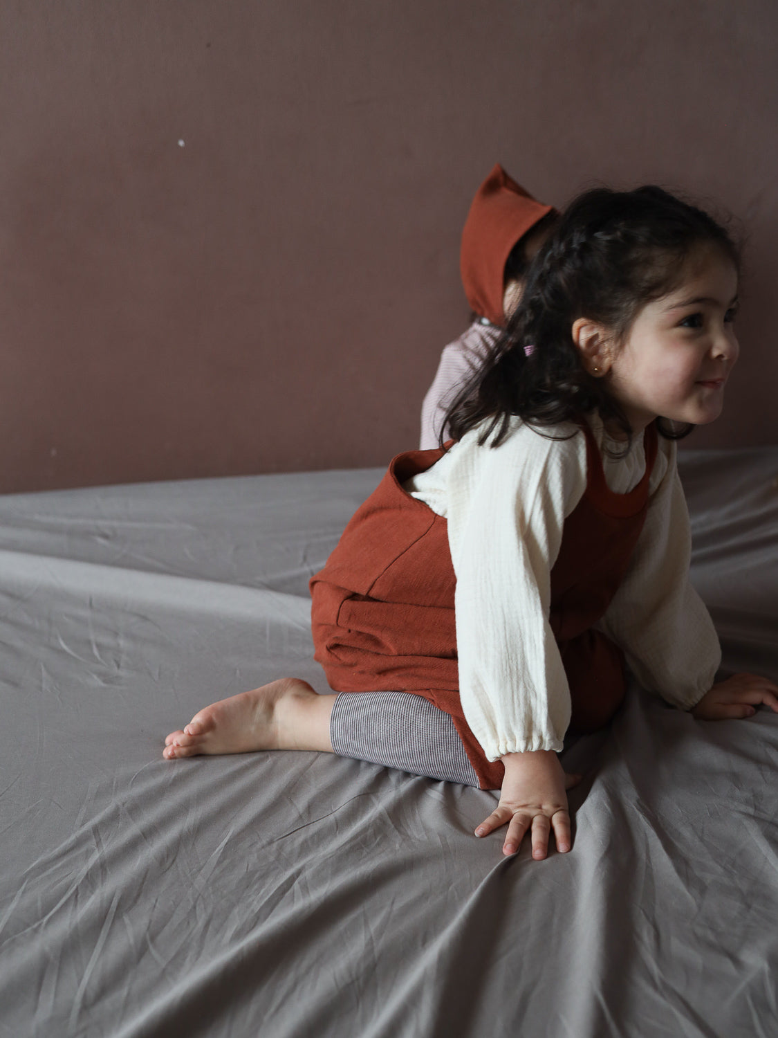 Kleinkind trägt Schlupflatzhose Leinen - Ziegel, kniet auf Bett, kombiniert mit cremefarbenem Oberteil und gestreifter Leggings. Einfache, bequeme Passform, perfekt für viel Bewegungsfreiheit.