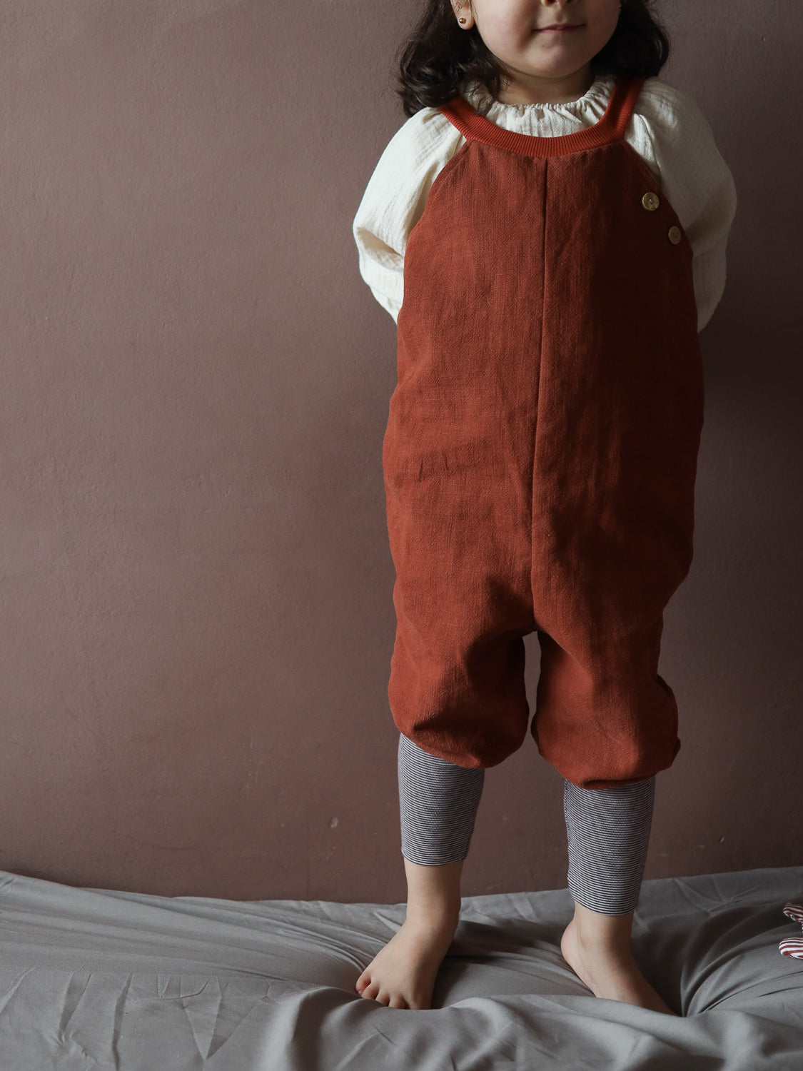 Kind in Schlupflatzhose Leinen - Ziegel, kombiniert mit gestreiften Leggings. Die rustfarbenen, lockeren Overalls mit Holztasten neben einem cremefarbenen Oberteil bieten Komfort und Bewegungsfreiheit auf weichem, grauen Untergrund.