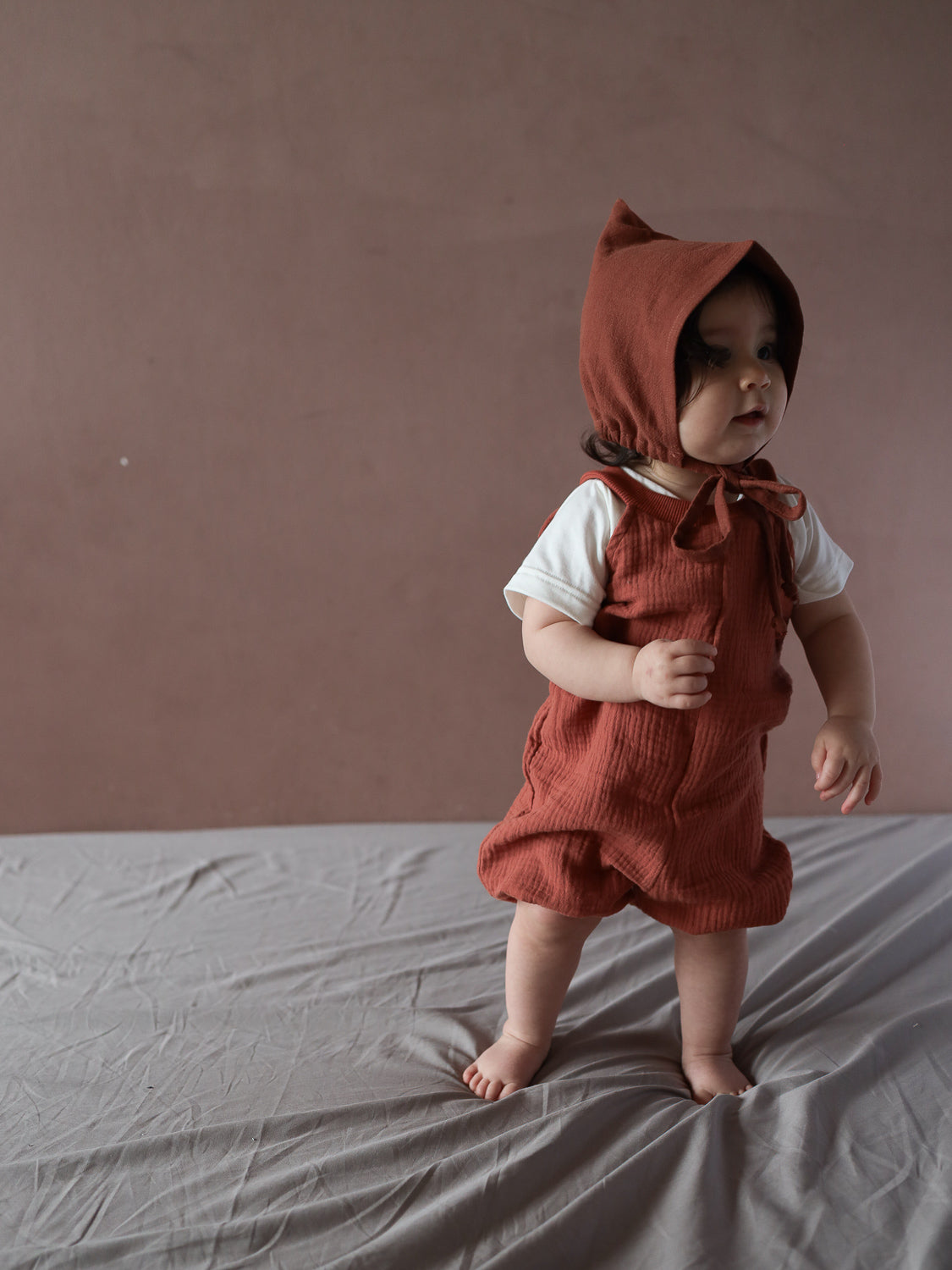 Ein Kleinkind trägt die Kurze Schlupflatzhose Bio-Musselin - Terrakotta, kombiniert mit einem passenden Bonnet und einem weißen T-Shirt, stehend auf einem weichen Stoff, mit erhobenen Händen für Balance.