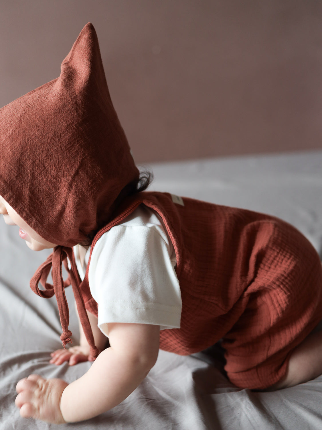 Ein Baby trägt die Kurze Schlupflatzhose Bio-Musselin - Terrakotta, mit passender Haube und weißem Shirt, krabbelt auf einer hellgrauen Oberfläche, zeigt den bequemen und praktischen Sommer-Einteiler.