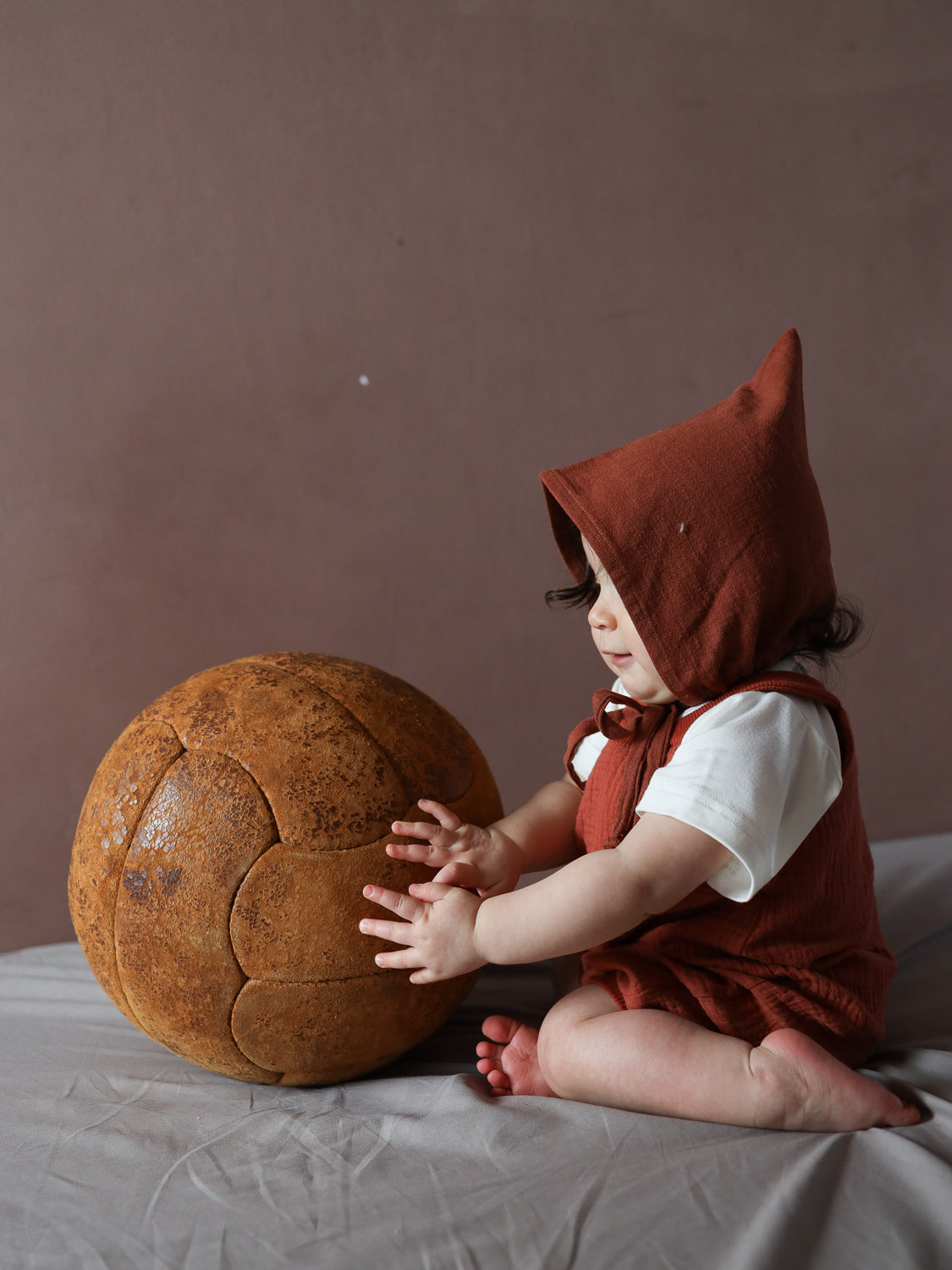 Ein kleines Kind trägt die Kurze Schlupflatzhose Bio-Musselin - Terrakotta über einem weißen Shirt und spielt mit einem großen, braunen Ball auf einer weichen, hellgrauen Unterlage.