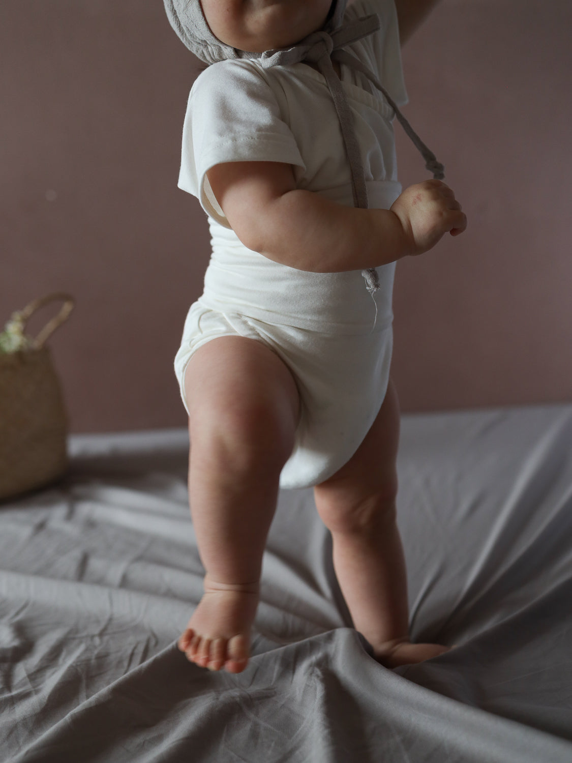 Ein Baby steht auf einem weichen Untergrund und trägt den Kurzarm Loopbody Bambusviskose - Natur, ein weißes, einteiliges Outfit mit kurzer Armform und weichem Stoff, ergänzt durch eine textile Mütze.