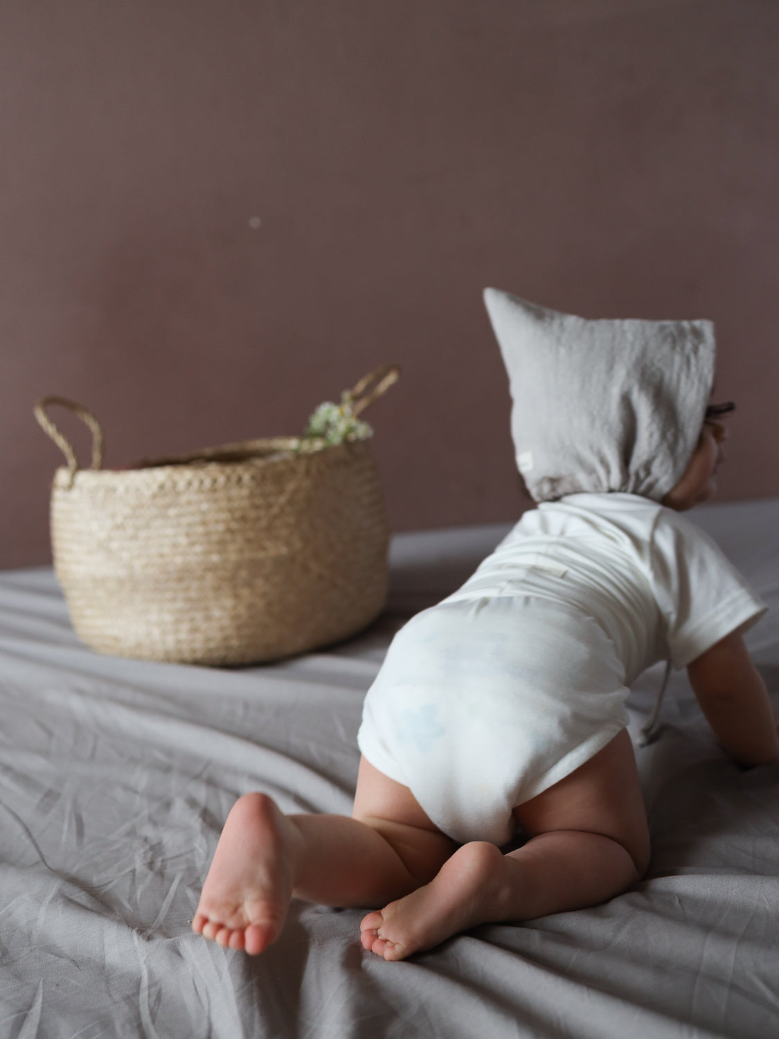 Ein Kleinkind in einem Kurzarm Loopbody aus Bambusviskose - Natur krabbelt über ein neutrales Bett. Es trägt einen grauen, spitzen Hut. Neben dem Bett steht ein geflochtener Korb.
