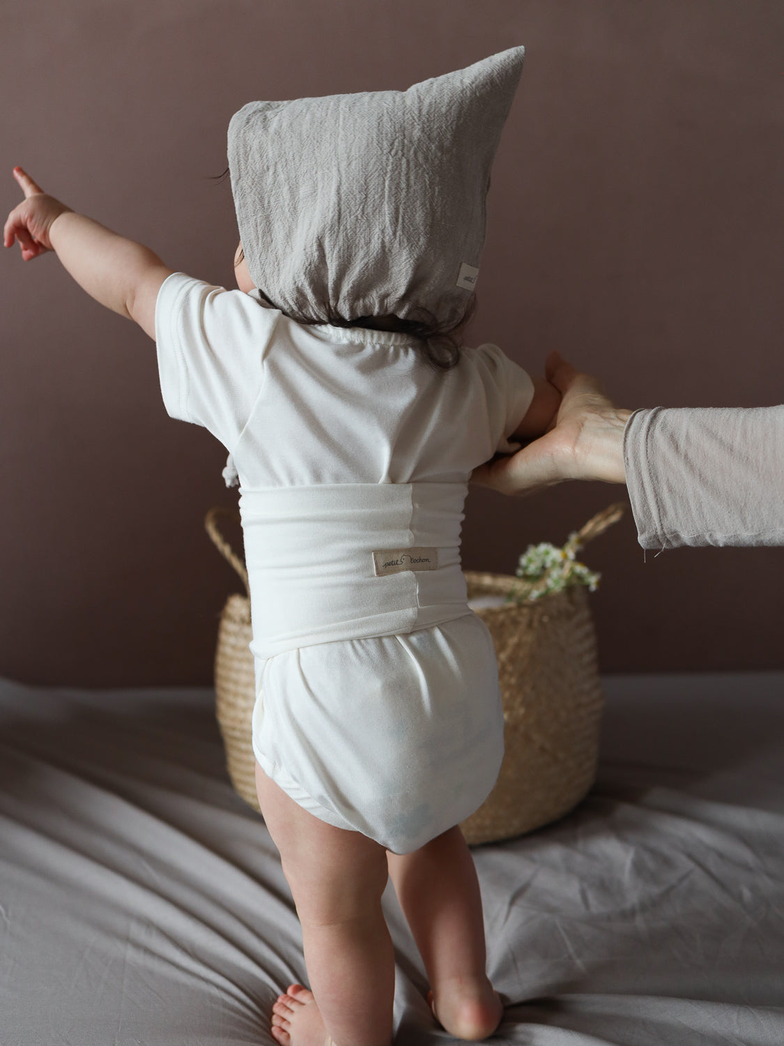 Ein Kleinkind trägt den Kurzarm Loopbody Bambusviskose - Natur, unterstützt von einer Hand. Der weiche Body umhüllt den Torso, ergänzt durch eine graue Kapuze, ideal für den Sommer.