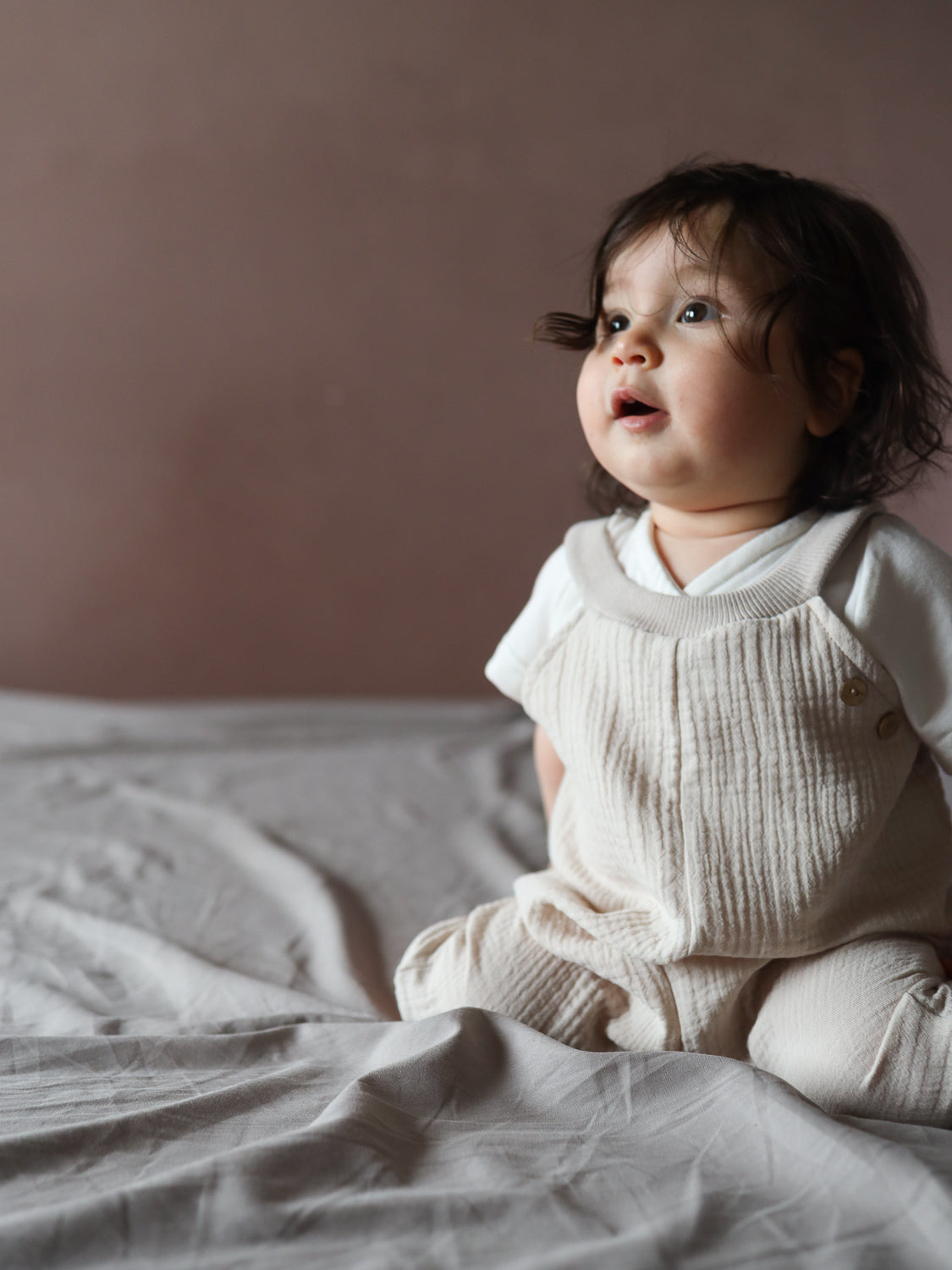 Ein Baby trägt eine Kurze Schlupflatzhose Musselin - Sand, handgenäht in Berlin, über einem weißen Shirt. Die komfortable, strukturierte Einteilerhose ist ideal für den Sommer und unterstützt Bewegungsfreiheit.