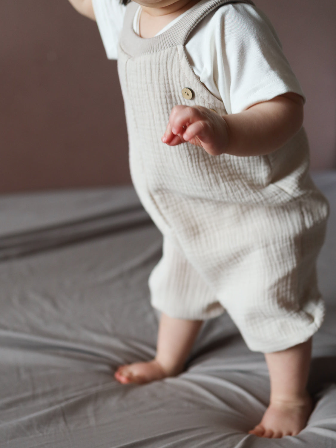 Ein Kleinkind trägt die Kurze Schlupflatzhose Musselin - Sand, einen kremfarbenen, strukturierten Einteiler, während es barfuß auf einem Bett steht und sich auf grauer Bettwäsche ausbalanciert.