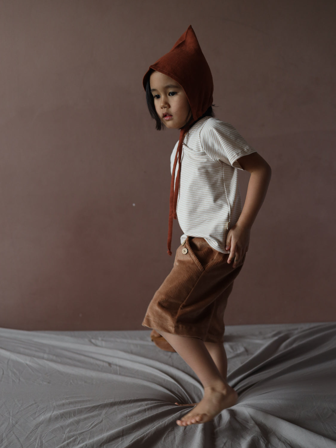 Ein Kleinkind trägt ein Kurzes Wickelshirt Baumwolle - Ringel Sand mit feinen Streifen, kombiniert mit braunen Cordshorts, während es auf einer weichen Unterlage steht.