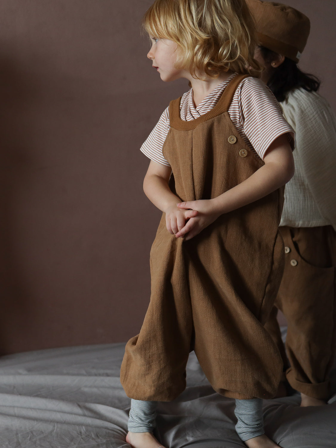 Ein Kind trägt das Kurzes Wickelshirt Baumwolle - Ringel Weizen, ein gestreiftes, kurzärmeliges Oberteil, kombiniert mit braunen Leinen-Overalls, auf weichem Untergrund vor schlichtem Hintergrund.