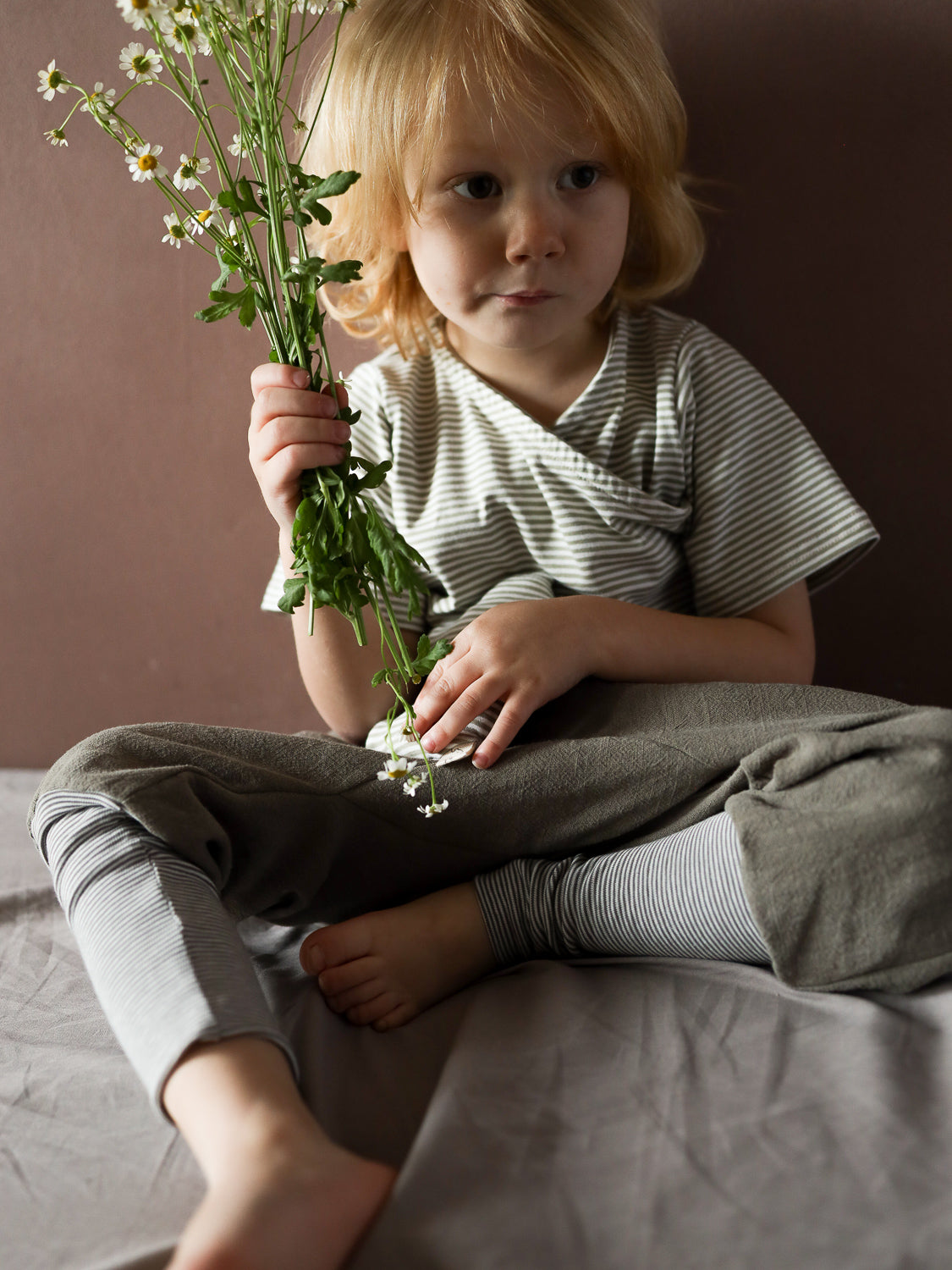 Ein Kind trägt das Kurzes Wickelshirt Baumwolle - Ringel Salbei, sitzt auf einem Bett und hält weiße Blumen. Das gestreifte Shirt ist weich und bequem, ideal für den Sommer.