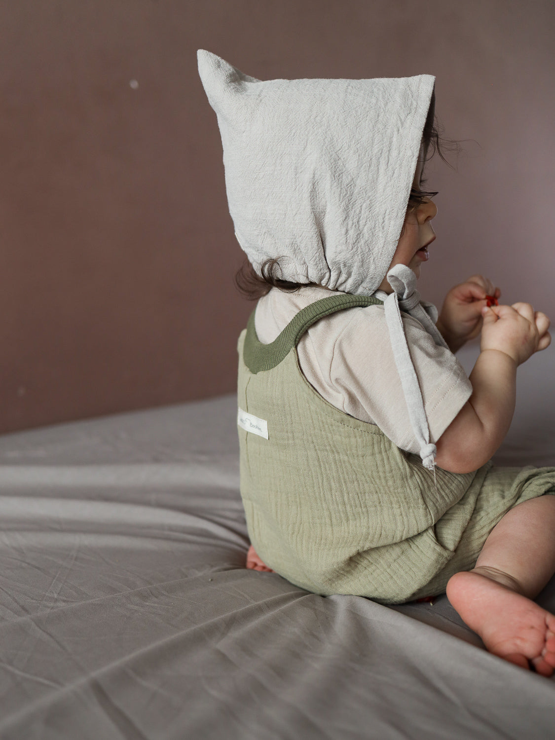 Ein kleines Kind trägt die Kurze Schlupflatzhose Bio-Musselin - Salbei, eine olivgrüne, handgenähte Musselin-Romper, über einem kurzärmeligen Shirt, während es auf einem Bett sitzt und ein rotes Objekt hält.