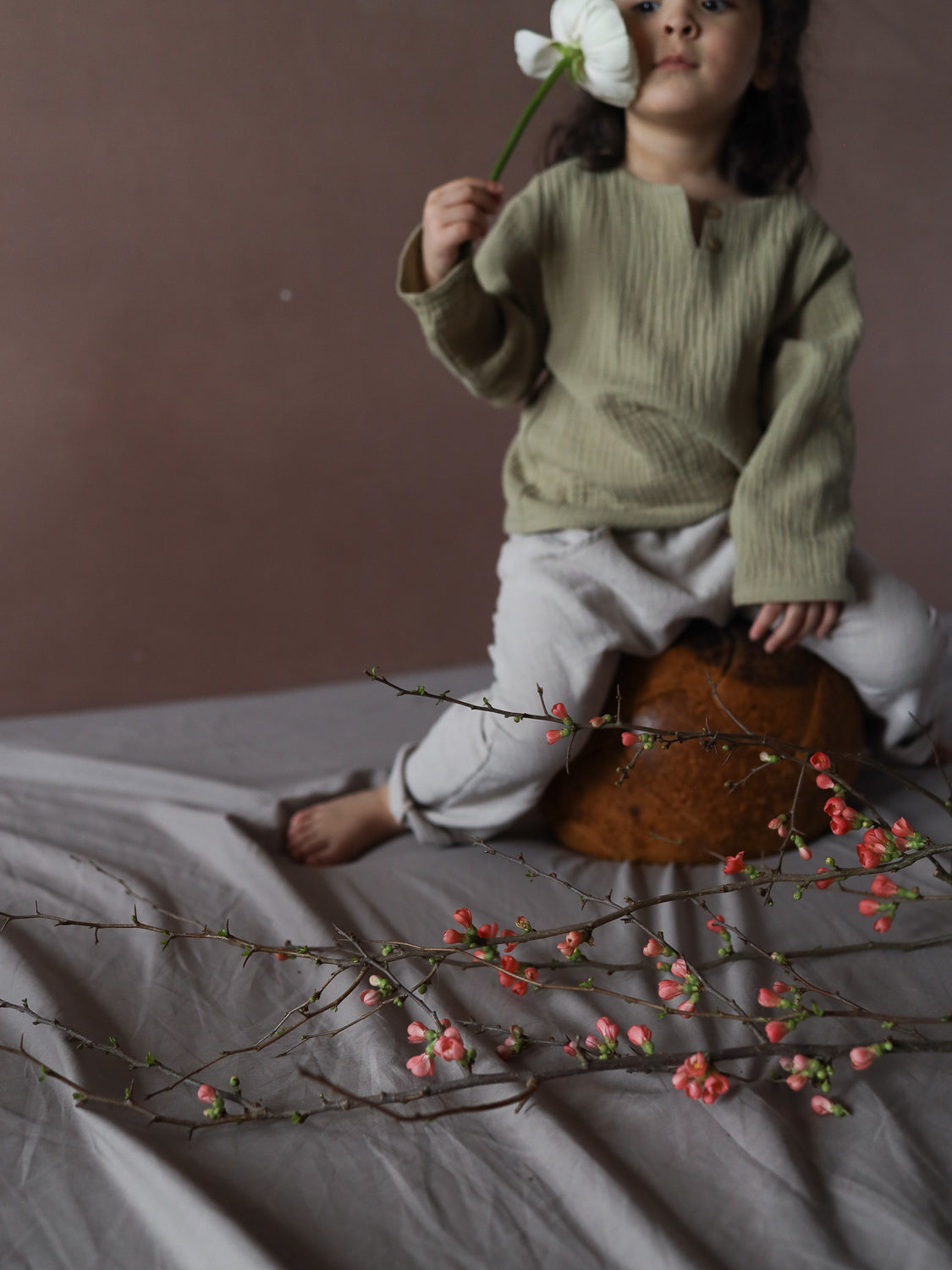 Kleines Kind auf russet-braunem Sitz, trägt Sommerknicker Leinen - Sand. Olivgrünes Oberteil, beige Hose, mit weißer Blume. Rosa Blütenzweige im Vordergrund, natürliche, bequeme Passform sichtbar.