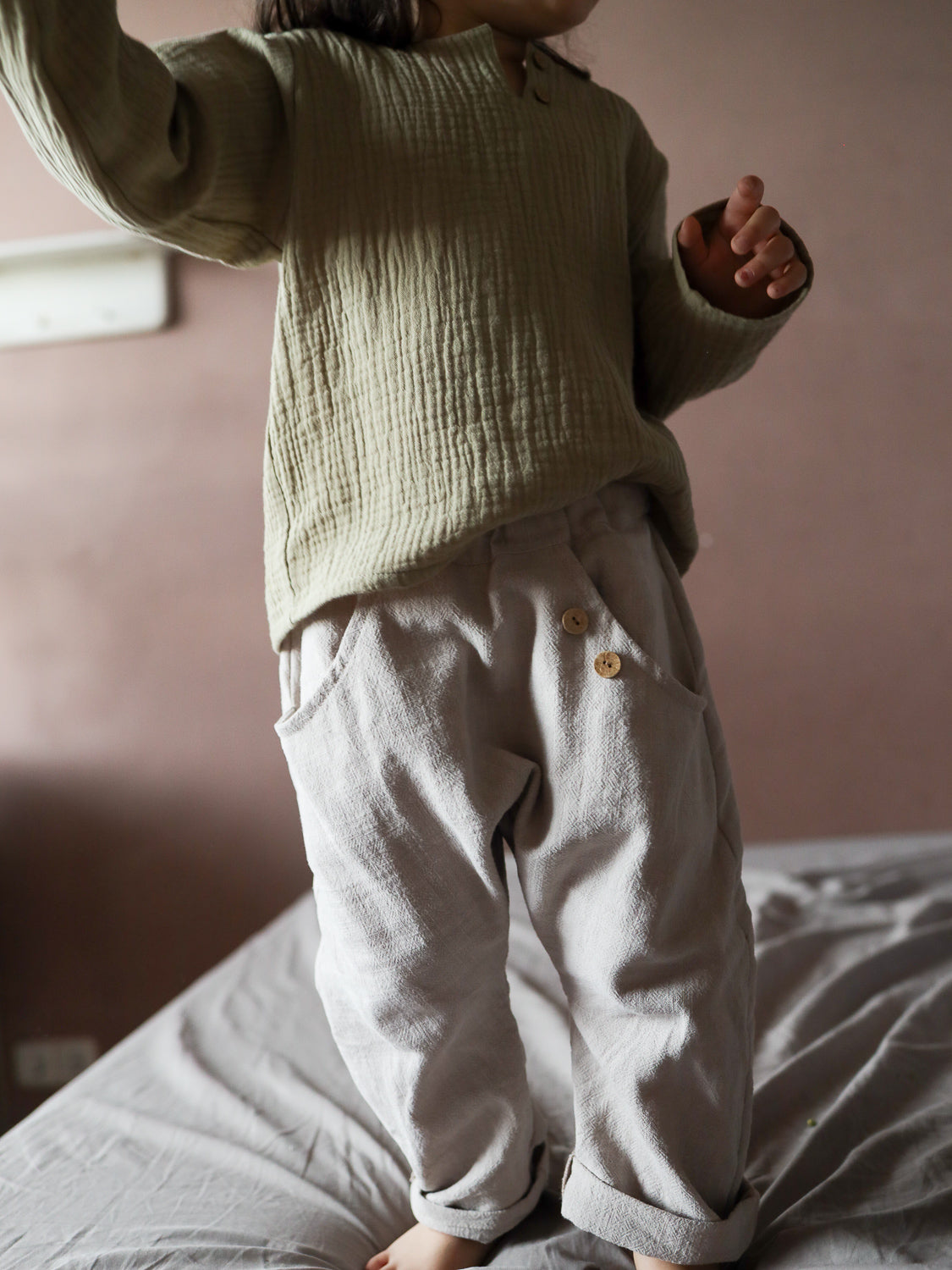Kind in neutraler Kleidung, trägt Sommerknicker Leinen - Sand: Locker sitzende, leichte Hose mit Holzknöpfen und Gummibund, ideal für Bewegungsfreiheit und Komfort. Perfekt für aktive Sommertage.