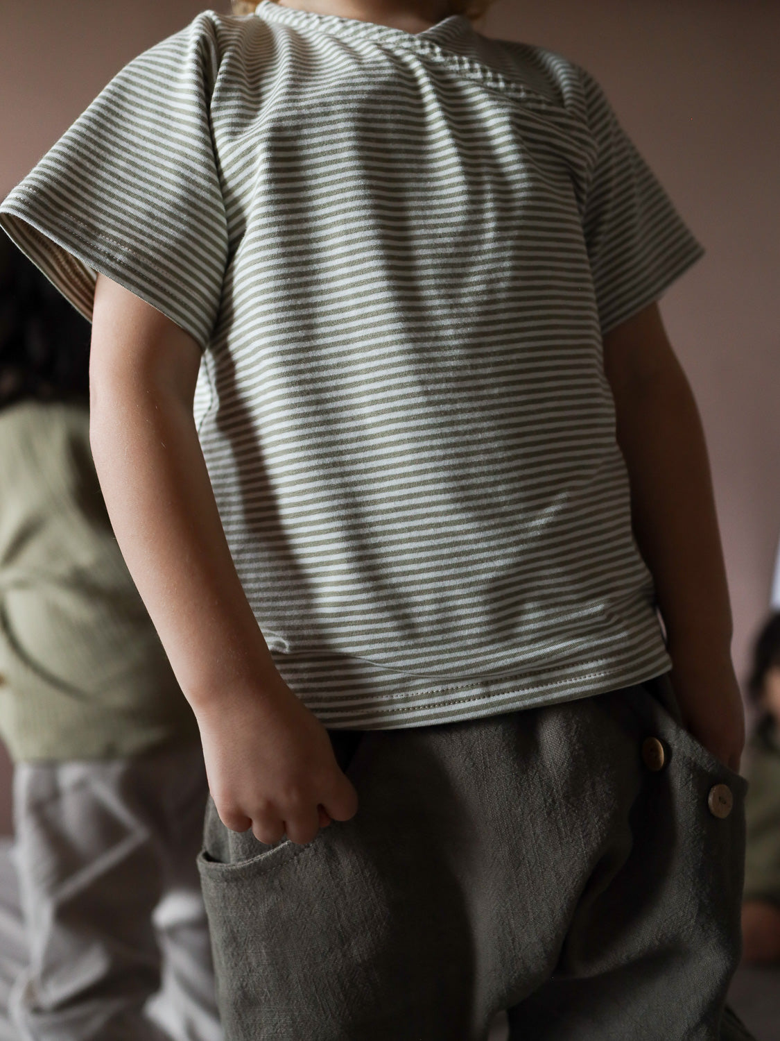 Zwei Kinder tragen das Kurzes Wickelshirt Baumwolle - Ringel Salbei. Im Vordergrund ein grünes, gestreiftes Shirt mit kurzen Ärmeln und Holzknöpfen an weiten, olivgrünen Hosen.