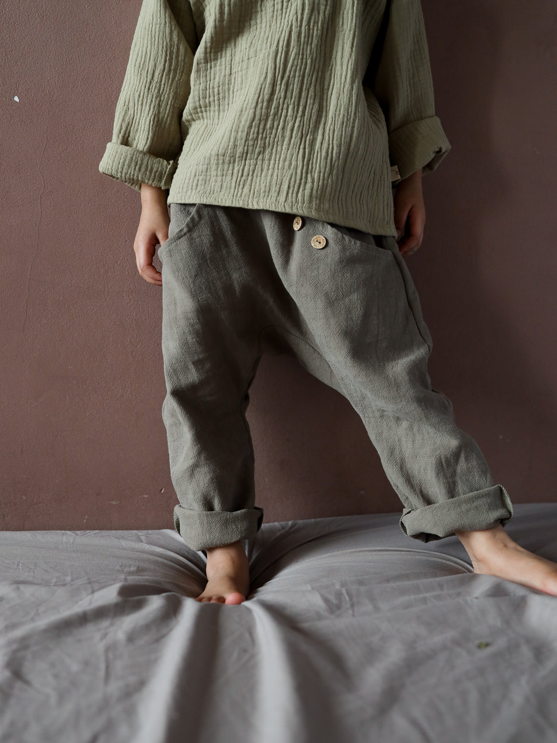 Kind steht barfuß auf grauem Kissen, trägt Sommerknicker Leinen - Oliv mit Holzknöpfen und lockerem Schnitt, ideal für Bewegungsfreiheit und Komfort, perfekt für aktive Sommertage.