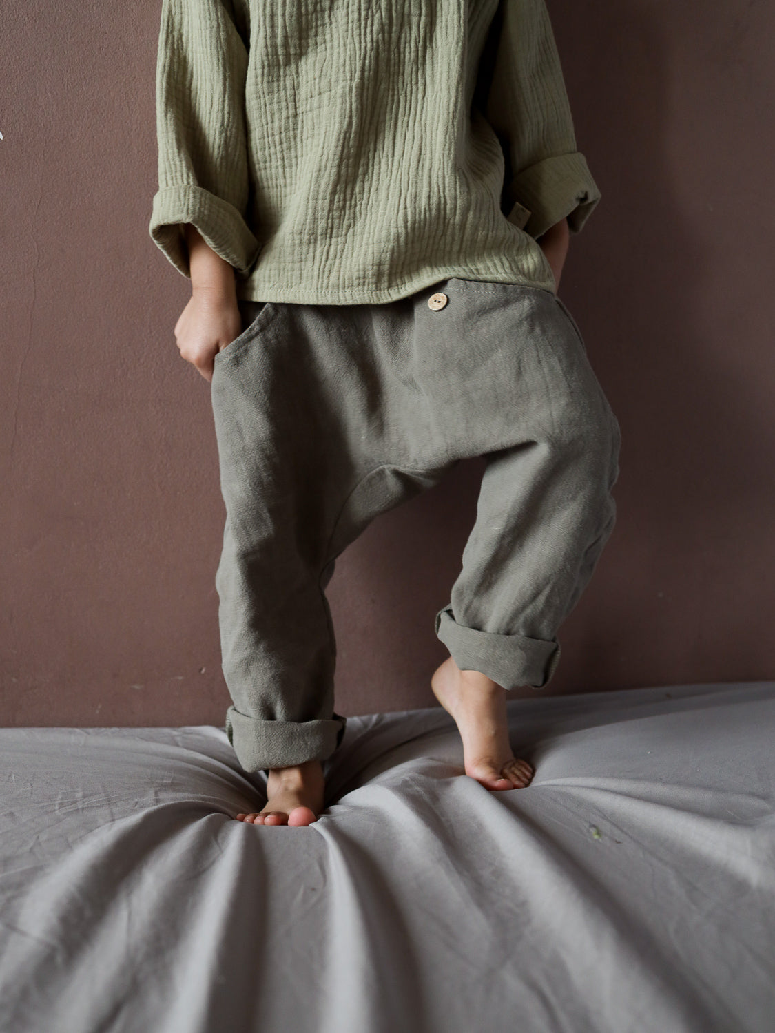 Sommerknicker Leinen - Oliv: Ein Kind trägt lockere, graugrüne Leinenhosen mit Holzknopf auf einer weichen Oberfläche. Die Hosen sind bequem, handgemacht und ideal für aktive Sommerabenteuer.
