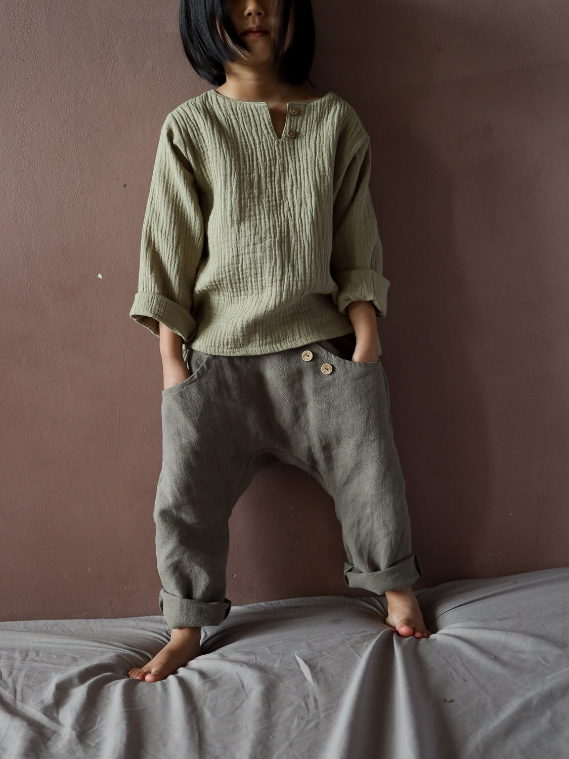 Kind trägt Sommerknicker Leinen - Oliv, eine lockere, graugrüne Hose mit Holzknöpfen, ideal für Bewegungsfreiheit. Perfekt handgemacht für den Sommer, kombiniert mit einem weichen, langärmeligen Oberteil.