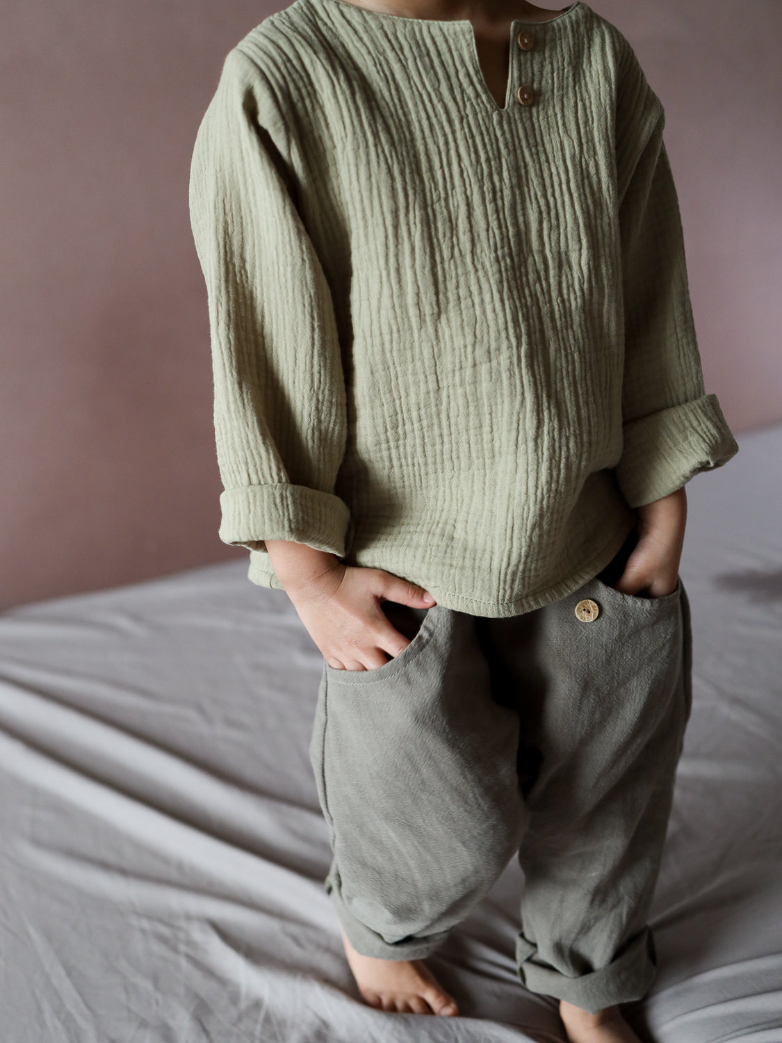 Ein Kind trägt die Sommerknicker Leinen - Oliv, eine locker sitzende Hose mit umgeschlagenen Bündchen und Knopfdetail, kombiniert mit einem langärmeligen, geknitterten Oberteil und Händen in den Hosentaschen.