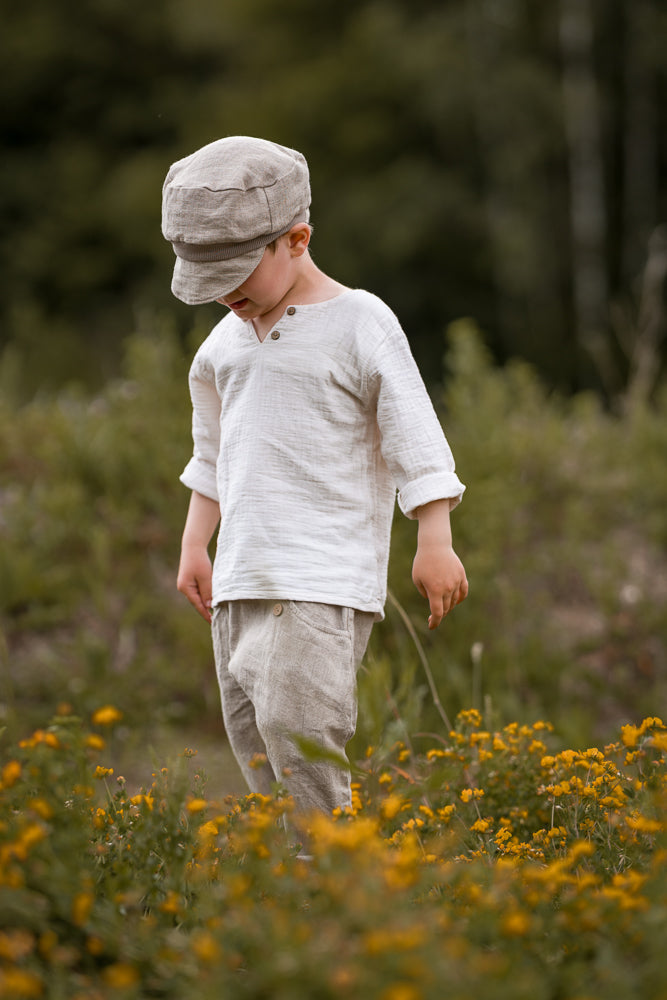 Ein Kind trägt die Sommerknicker Leinen - Sand, bestehend aus lockerem, langärmligem Leinenhemd und passender Hose. Der weiche Gummibund sorgt für Komfort, ideal für aktive Sommertage.