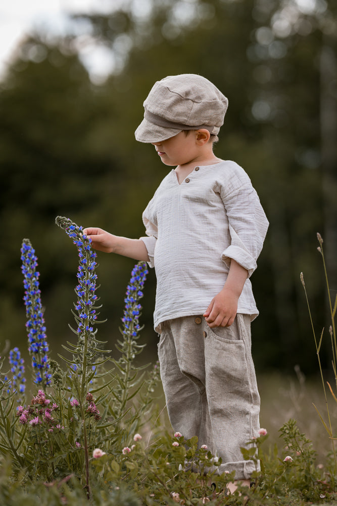 Ein Kind trägt Sommerknicker Leinen - Sand, inspiziert blau blühende Lupinen. Die luftige, handgemachte Leinenhose ist locker geschnitten mit einem bequemen Gummibund, ideal für aktive Tage im Freien.