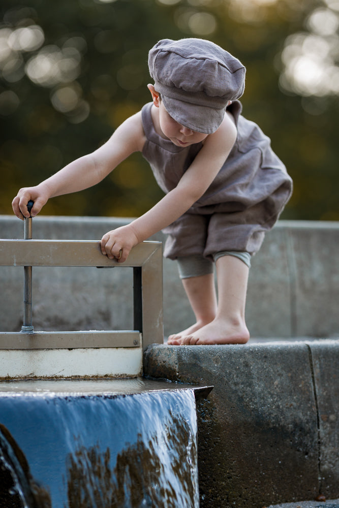 Kleinkind in Schlupflatzhose Leinen - Oliv, steht barfuß an einem Wasserbecken und dreht einen kleinen Metallgriff. Trägt passende Schiebermütze, zeigt Bewegungsfreiheit und natürliche Eleganz der Hose.