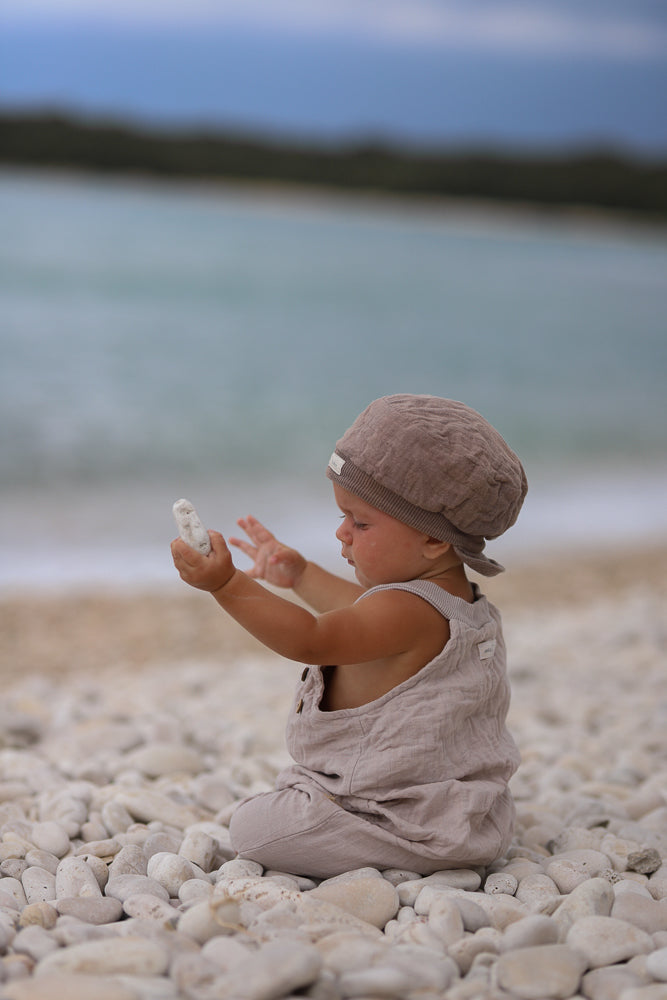 Ein Kleinkind in einer Kurze Schlupflatzhose Bio-Musselin - Sand sitzt am Kiesstrand, betrachtet einen weißen Stein. Das Outfit besteht aus weichem Leinen und einer passenden Mütze.