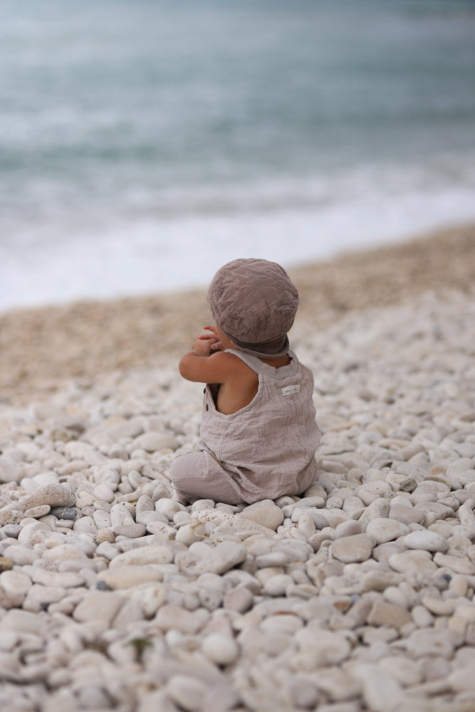Kleinkind am Strand, trägt eine Kurze Schlupflatzhose Bio-Musselin - Sand, zeigt Rückenansicht. Sommer-Einteiler aus weichem Stoff, ideal für Bewegungsfreiheit, in Berlin handgenäht, umgeben von Kieselsteinen und Meer.