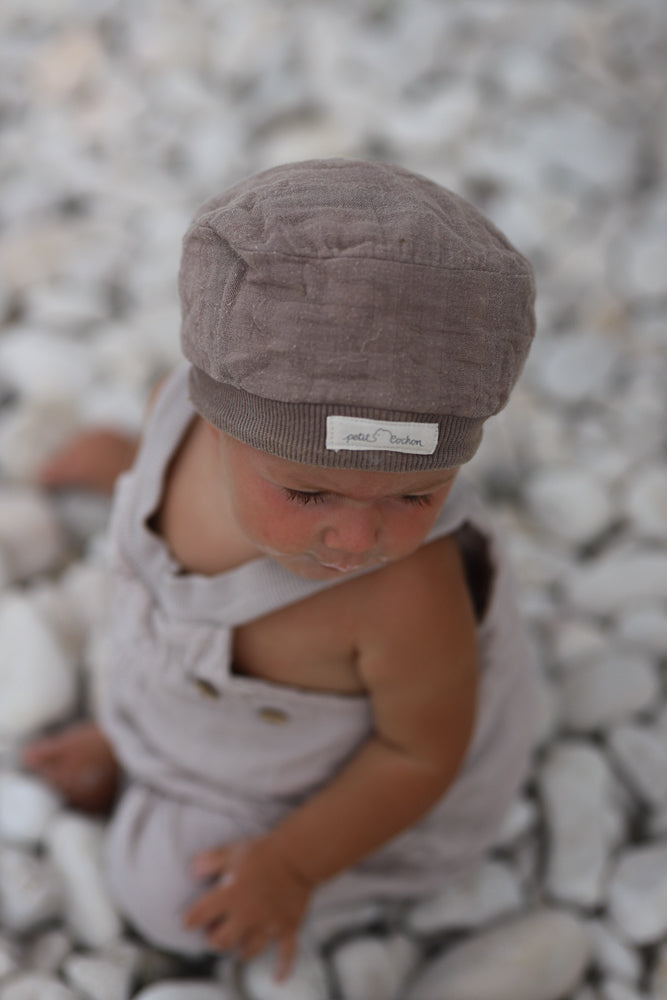 Ein Baby in neutralen Leinenoveralls sitzt auf Steinen. Die kurze Schlupflatzhose Bio-Musselin - Sand ist bequem, handgenäht in Berlin, perfekt für sommerliche Abenteuer.