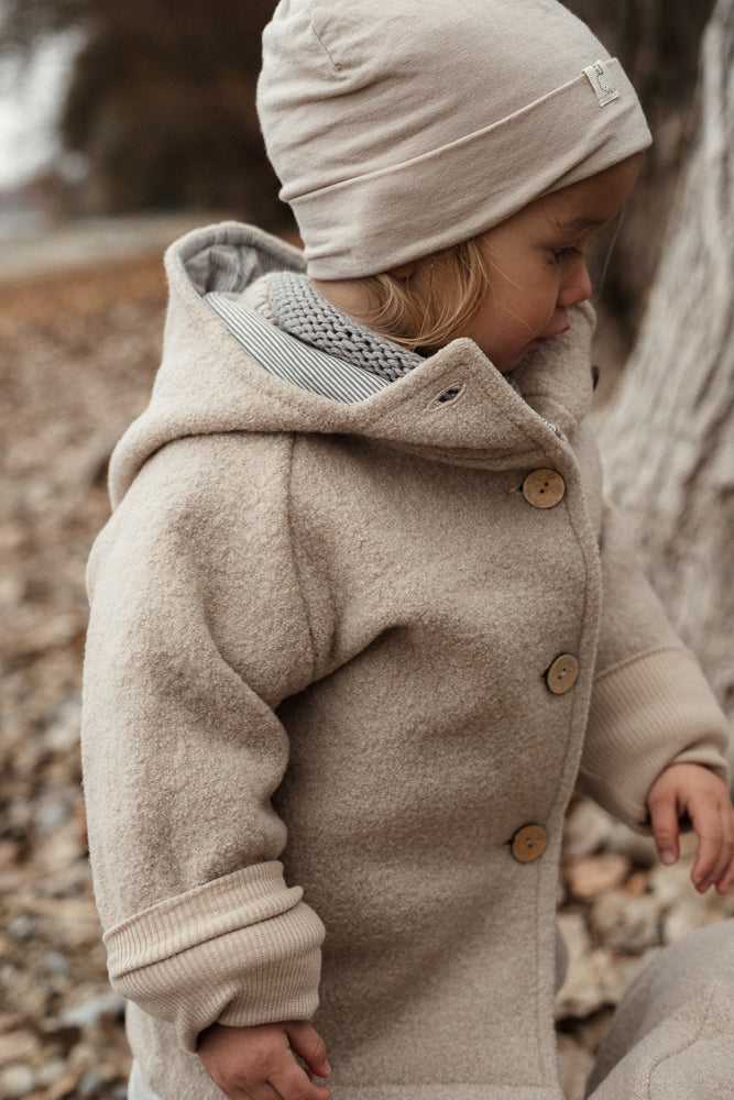 Ein Kind im Freien trägt die Wichteljacke Wollwalk - Sand mit Holzknöpfen und passender Mütze, ideal für Herbst. Die Jacke bietet Bewegungsfreiheit und temperaturregulierende Eigenschaften.