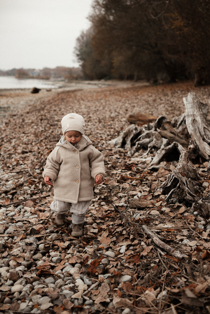 Ein kleines Kind trägt die Wichteljacke Wollwalk - Sand, spaziert am kies- und blattbedeckten Ufer entlang. Die beige Jacke und der passende Hut bieten Schutz und Komfort bei kühlem Wetter.