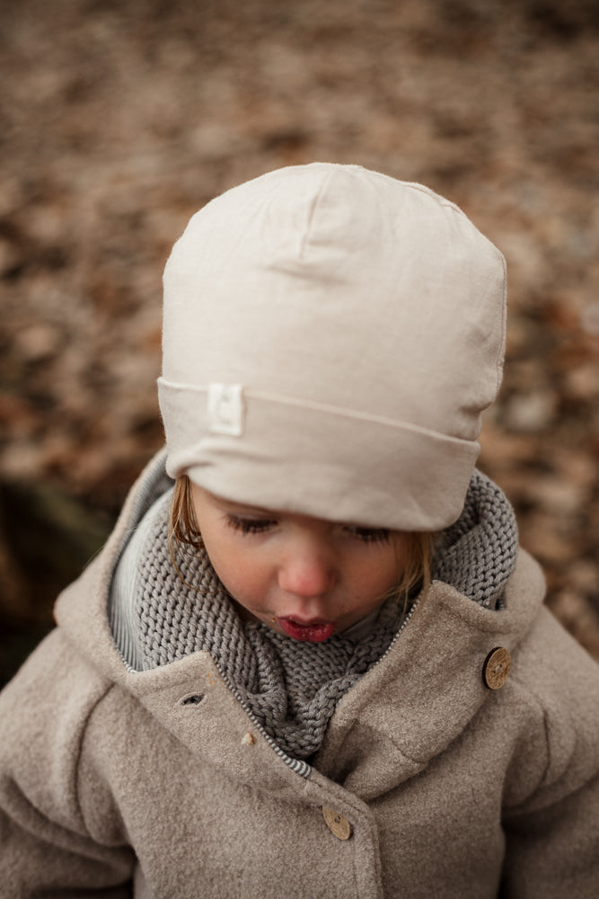 Ein Kind trägt die Wichteljacke Wollwalk - Sand mit Holzknöpfen, kombiniert mit einem cremefarbenen Beanie und einem Strickschal, perfekt für kühle Herbst- und Wintertage.