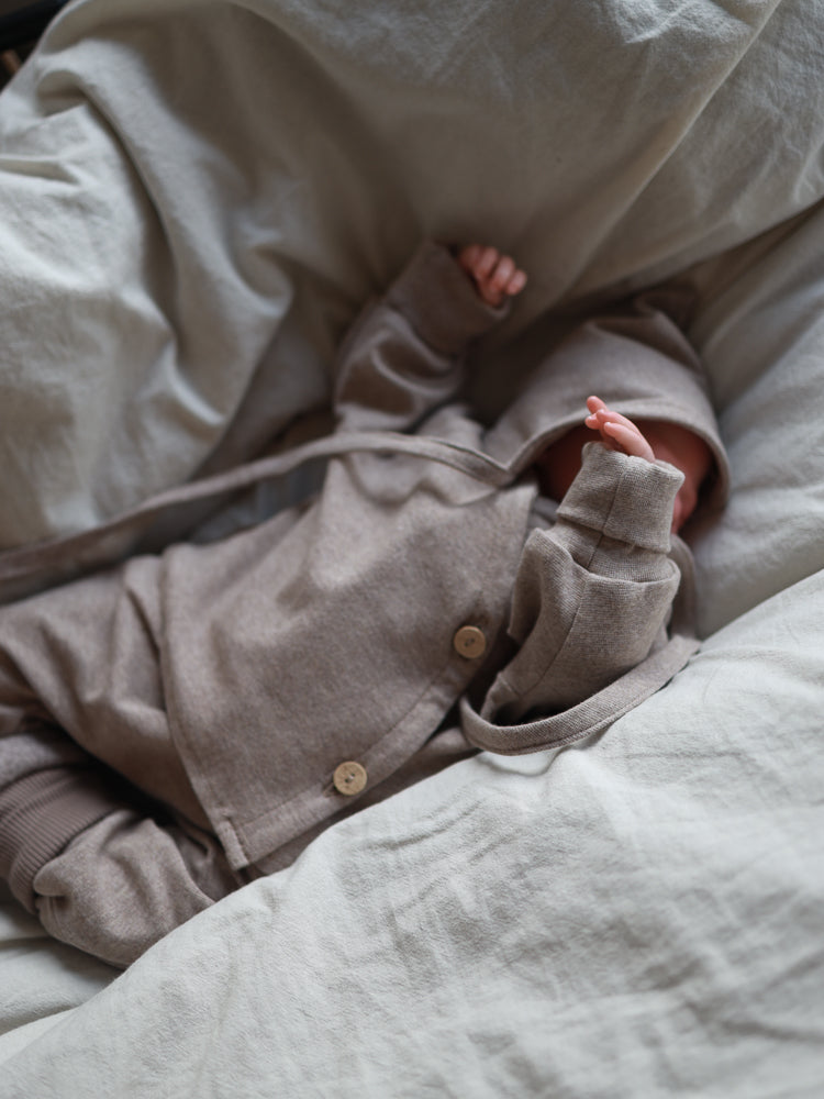 Neugeborenes mit Zwergenmütze Feinstrick - Hafer Meliert, warmes, weiches Jersey mit Zipfel, liegt auf neutralem Bett, Outfit beige, ideal für empfindliche Babyhaut, stilvoll und funktional in klassischer Zwergenform.