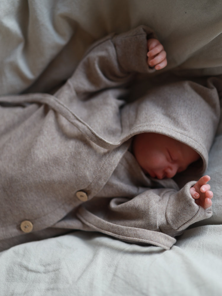 Ein Neugeborenes schläft in einer Zwergenmütze Feinstrick - Hafer Meliert. Die weiche, graue, melierte Feinstrick-Jersey-Mütze bedeckt den Kopf, während kleine Hände und das Gesicht teilweise sichtbar sind.