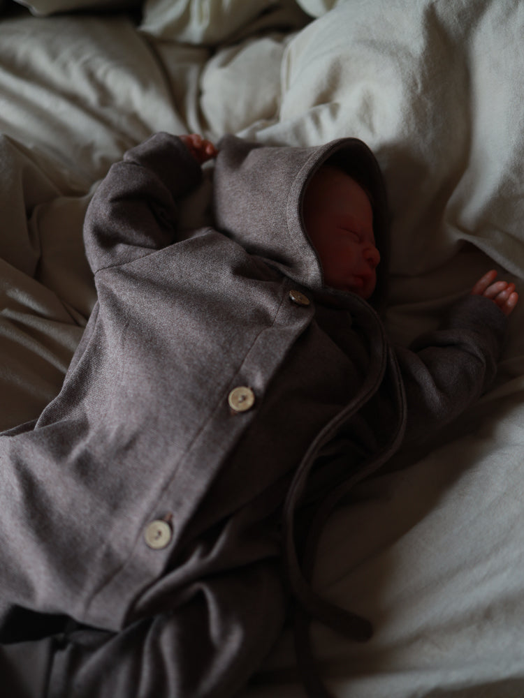 Neugeborenes in einem Mini Overall Feinstrick - Praline Meliert, schlafend auf hellem Untergrund. Der Overall hat lange Ärmel, Knöpfe und eine Kapuze, bietet Bewegungsfreiheit und weichen Tragekomfort.