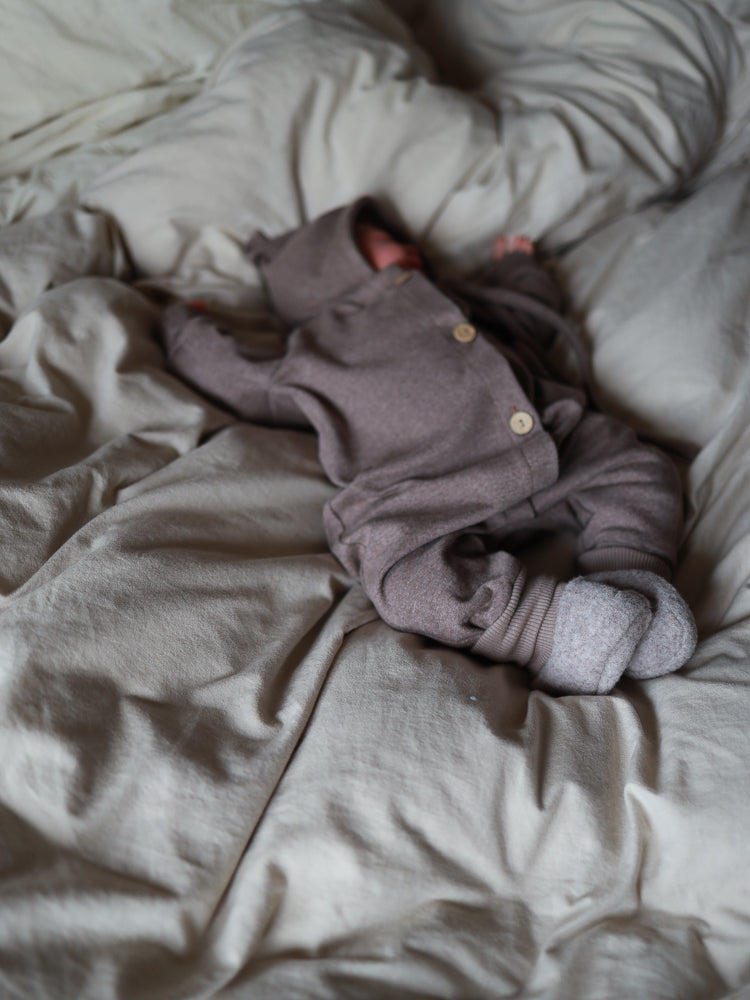 Ein Baby liegt auf einer weichen Decke, gekleidet im Mini Overall Feinstrick - Praline Meliert, mit Kapuze und passenden Stoffstiefeln, ideal zum Schlafen oder Spielen, hergestellt in Berlin.