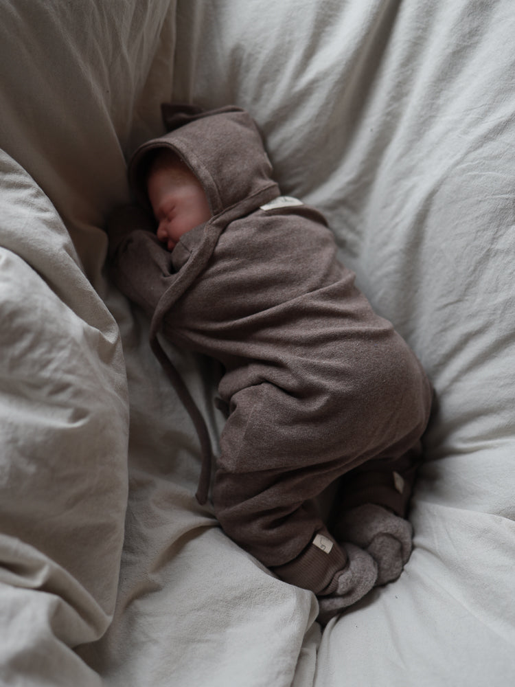 Ein schlafendes Neugeborenes trägt den Mini Overall Feinstrick - Praline Meliert, einen warmen, melierten braunen Einteiler aus elastischem Feinstrick-Jersey, der Bewegungsfreiheit und Komfort bietet.