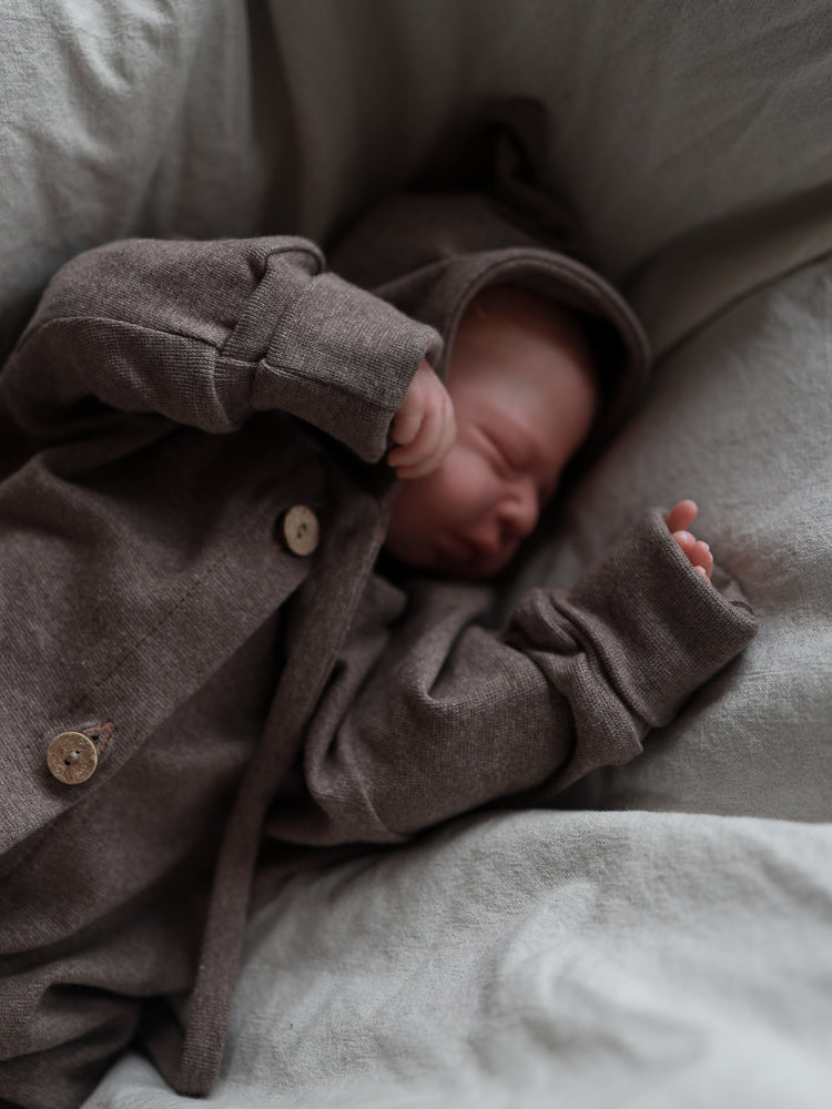 Neugeborenes in einem Mini Overall Feinstrick - Praline Meliert mit Kapuze und Holzknöpfen, schläft friedlich auf weichem Untergrund. Der Overall bietet elastischen Komfort und Bewegungsfreiheit.