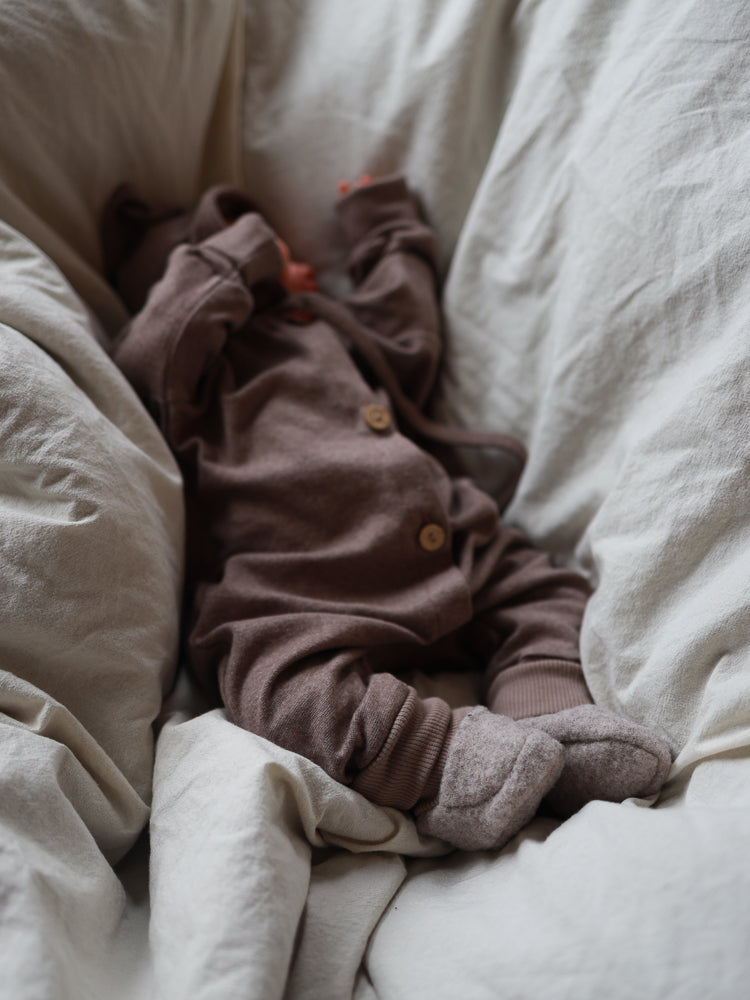 Ein Baby liegt auf cremefarbenem Bettzeug, trägt einen braunen Strampler und dazu passende Babybooties Merino Wollfleece - Hafer, die kleine Füße warm und bequem halten.