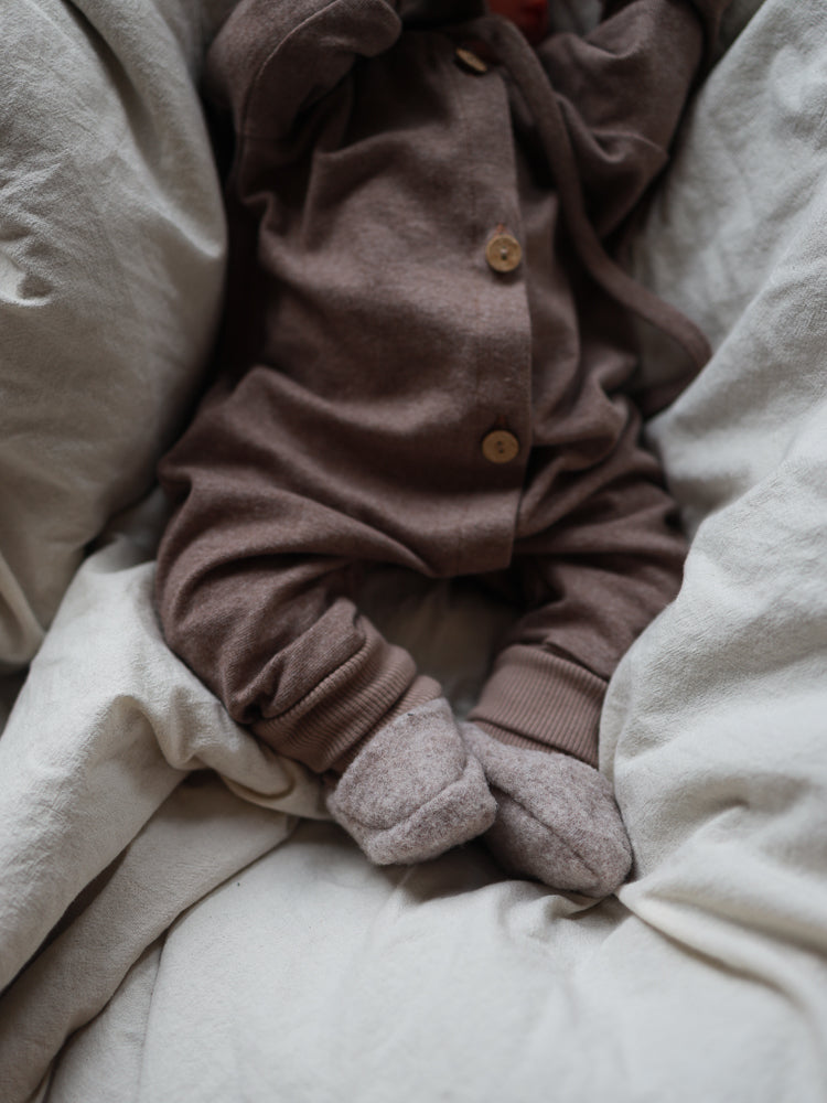 Der Mini Overall Feinstrick - Praline Meliert zeigt ein Baby auf einer weichen Decke, gekleidet in einem braunen, melierten, elastischen Strickanzug mit passenden Fußhandschuhen, ideal für Komfort und Bewegungsfreiheit.