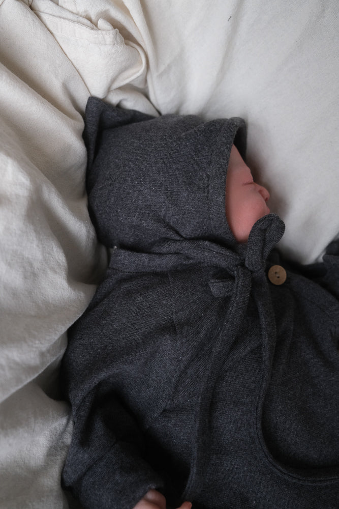 Ein schlafendes Baby trägt die Zwergenmütze Feinstrick - Ruß Meliert, aus weichem, atmungsaktivem Jersey, mit Kapuze und Schleife am Hals, auf hellem Bettzeug liegend.