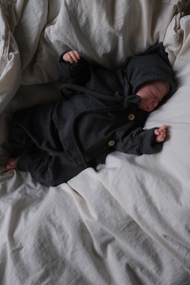 Neugeborenes schläft in einem dunkelgrauen, kapuzenartigen Zwergenmütze Feinstrick - Ruß Meliert auf weichem Bett, Arme ausgestreckt, bekleidet mit meliertem Jersey mit Holzknöpfen, modern und komfortabel gefertigt.