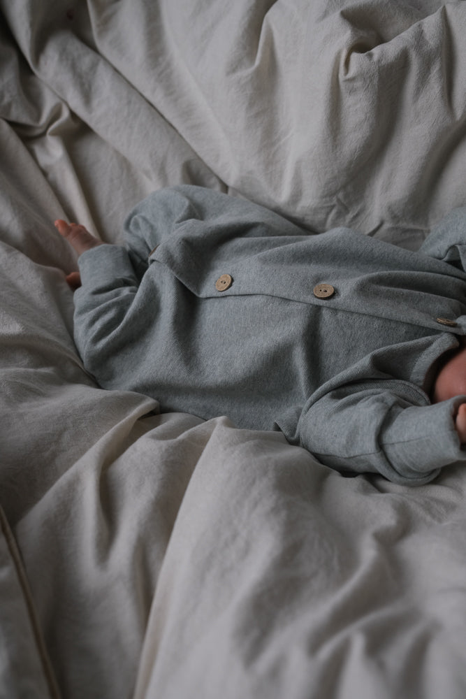 Baby im Mini Overall Feinstrick - Mint Meliert liegt entspannt auf weicher, beiger Bettwäsche. Das Langarm-Design mit Holzknöpfen bietet Komfort und Bewegungsfreiheit für Spielen oder Schlafen.
