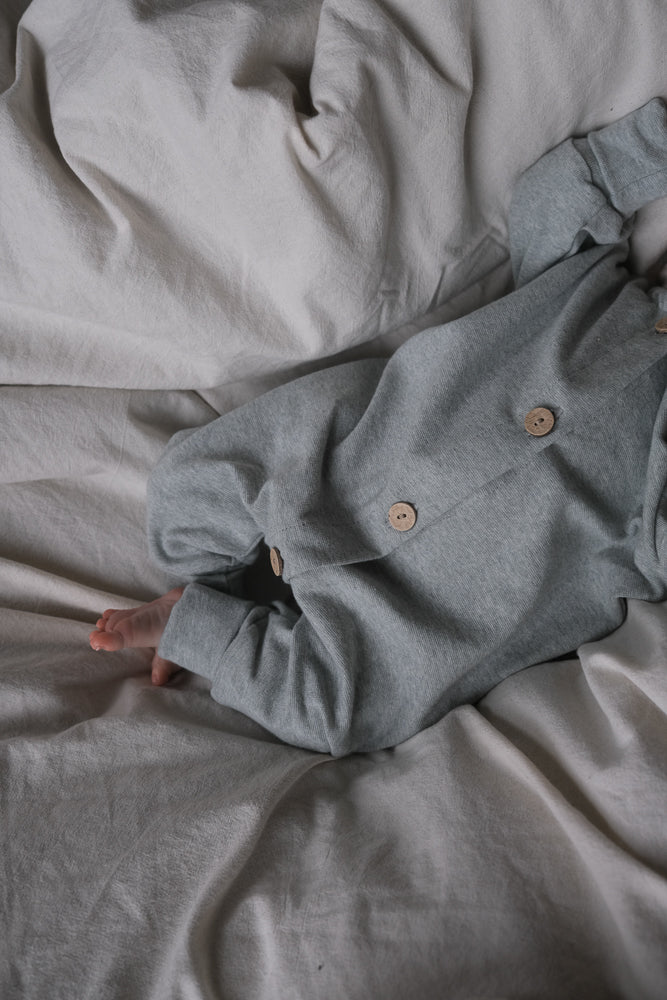 Kleinkind liegt auf zerknitterter Bettwäsche, trägt Mini Overall Feinstrick - Mint Meliert mit grauem Jersey, Holzknöpfen und zeigt einen nackten Fuß für Bewegungsfreiheit und Komfort.