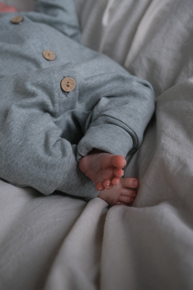 Mini Overall Feinstrick - Mint Meliert: Ein Baby liegt auf einer weichen Unterlage, trägt einen grauen Langarm-Overall mit Holzknöpfen vorne; winzige Füße sind sichtbar.