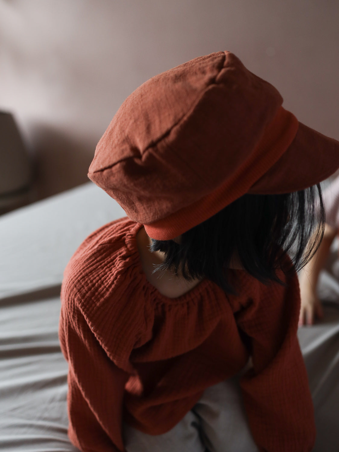 Ein Kind sitzt auf einem Bett, trägt ein rust-orangefarbenes, strukturiertes Oberteil und die Michelmütze Leinen - Ziegel, mit dunklem Haar, das unter der weichen Mütze hervorschaut.