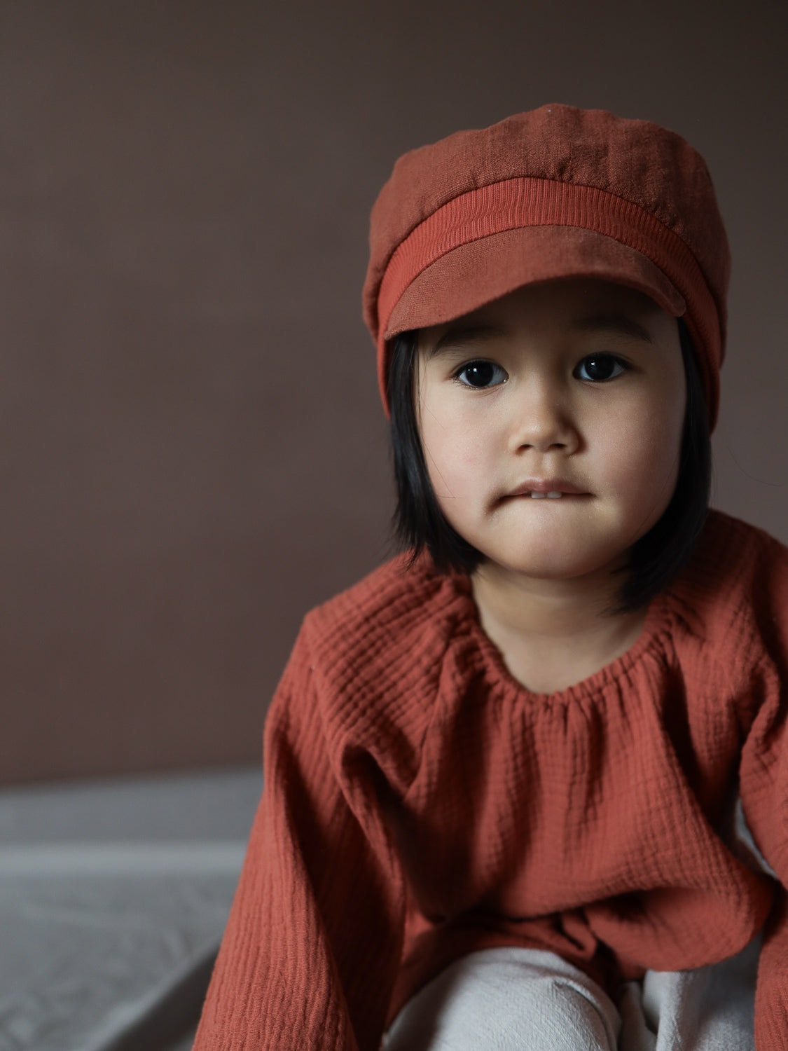 Ein Kleinkind trägt die Michelmütze Leinen - Ziegel, passend zu einem strukturierten Langarmshirt, mit nachdenklichem Blick und leichtem Lippenbeißen, während es leicht abseits der Kamera schaut.