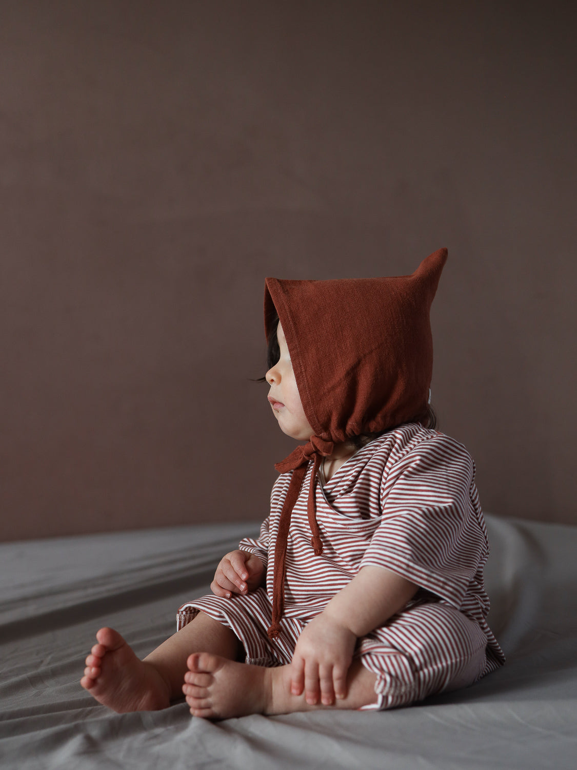 Ein Kleinkind trägt die Zwergenmütze Leinen - Ziegel, eine spitz zulaufende, gebundene Mütze, kombiniert mit einem rot-weiß gestreiften Outfit. Die Mütze bietet Schutz und Komfort.