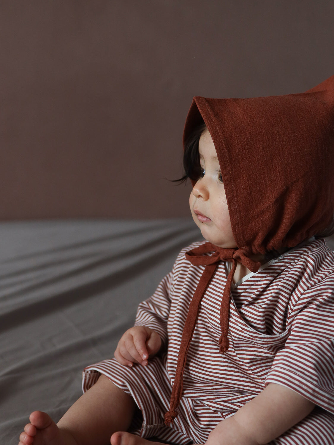 Kleinkind in Zwergenmütze Leinen - Ziegel trägt rustbraune Haube und gestreiften Einteiler; sitzt mit seitlichem Blick, zeigt Fuß und Hand auf Knie, umrahmt von warmen Erdtönen.