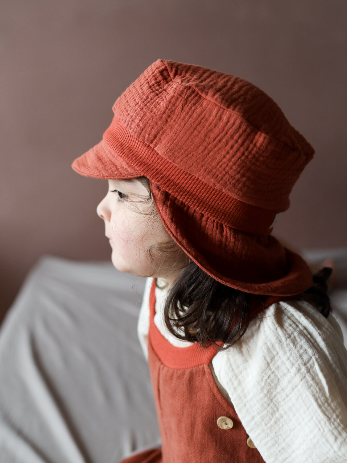 Ein Kind trägt die Michelmütze Bio-Musselin mit Nackenschutz - Terrakotta, eine luftige, strukturierte Sommermütze mit Ohrenklappen und elastischem Bündchen, kombiniert mit einem passenden roten Kleid und cremefarbenem Oberteil.