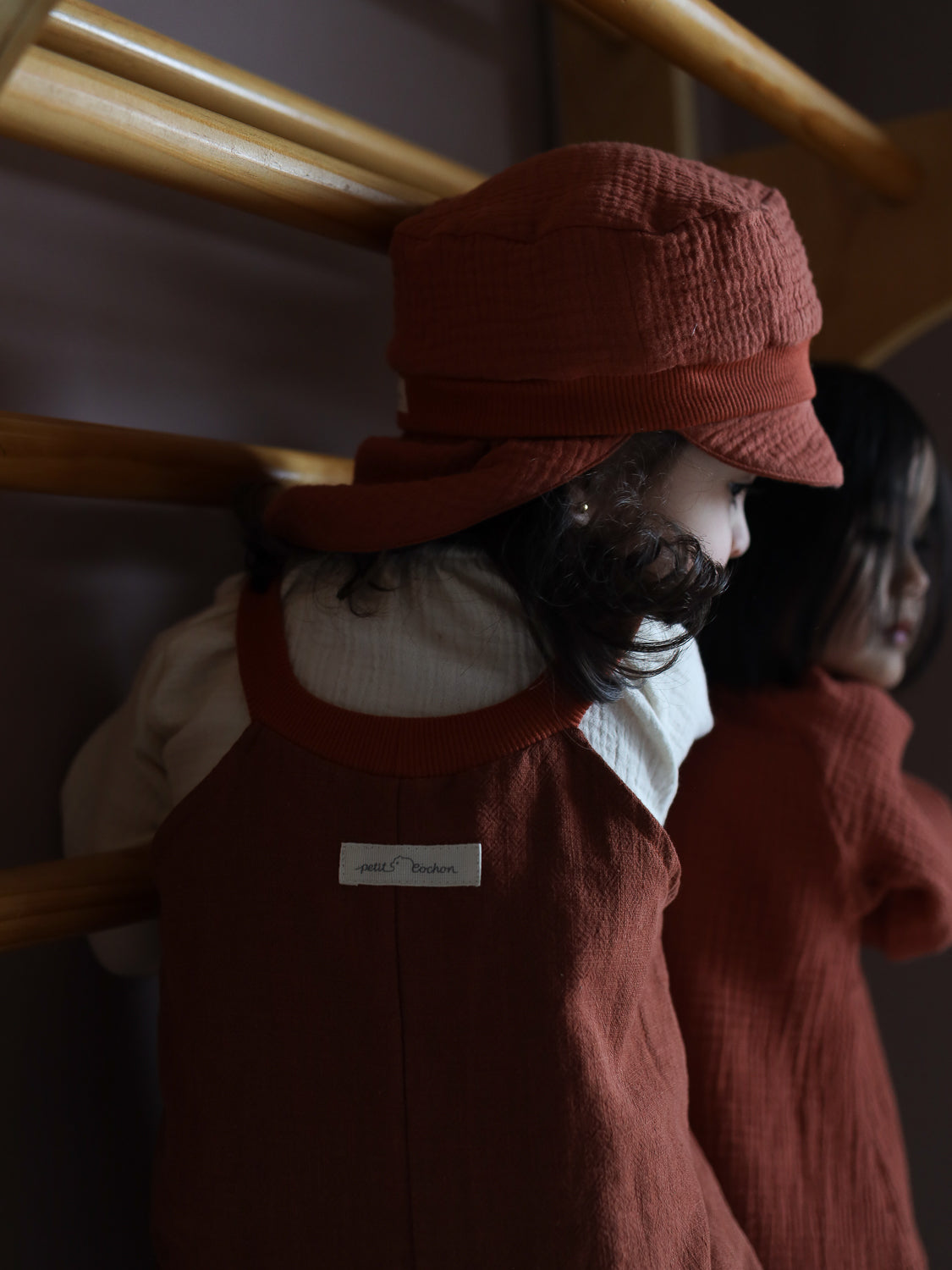 Ein Kleinkind trägt die Michelmütze Bio-Musselin mit Nackenschutz - Terrakotta aus Bio-Baumwolle, kombiniert mit passendem Outfit und steht vor Holzspielstangen, was eine warme, intime Atmosphäre schafft.